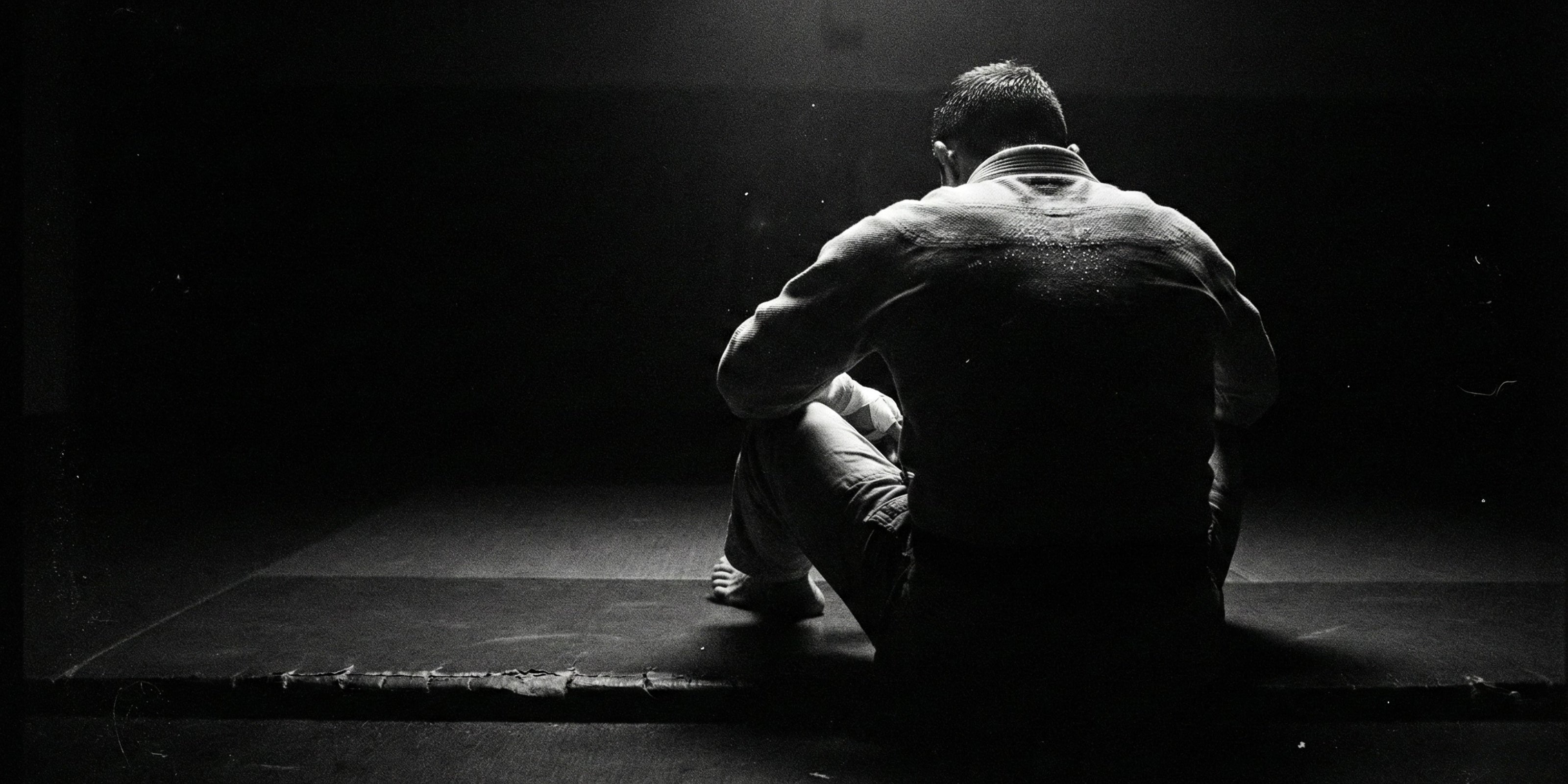 Desktop Header: BJJ Athlet im Gi sitzt fokussiert auf der Matte. Dramatische Schwarz-Weiß Ästhetik visualisiert das Mindset und die Ruhe nach dem Kampf.