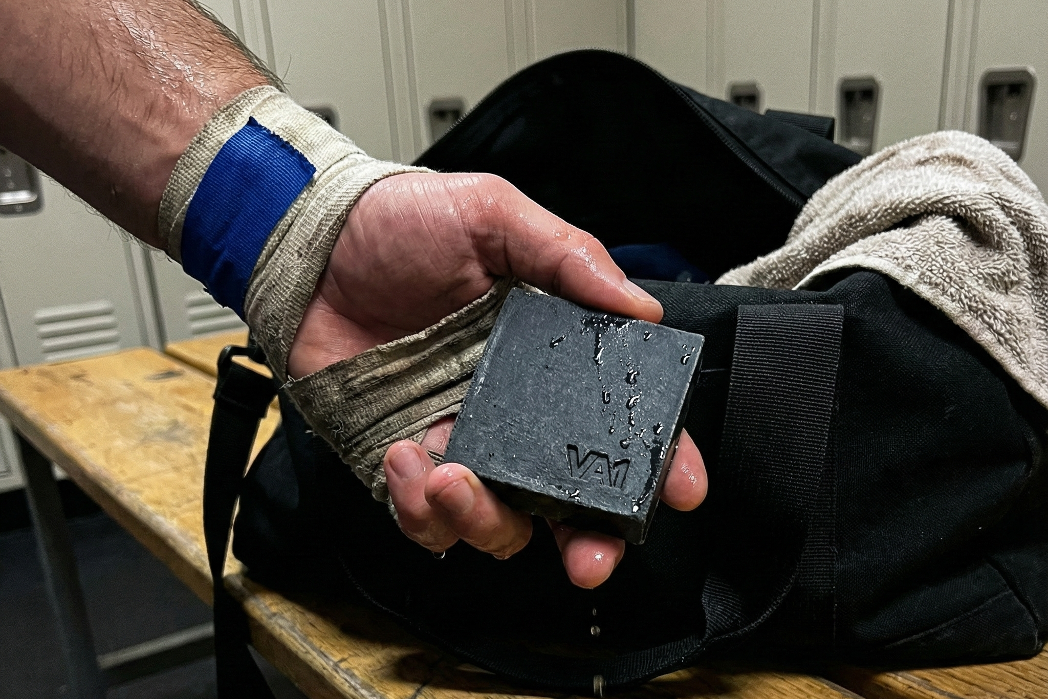 Bandagierte Hand mit blauem Tape hält nasse VA1 Core Bar über der Sporttasche. Aftermath Hygiene Routine direkt nach dem Training.